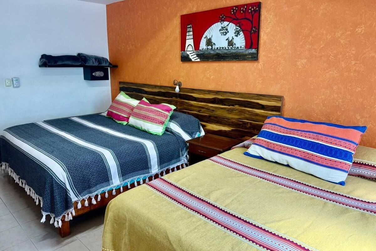 A room with two beds, each with a different patterned bedspread. There is a wall-mounted shelf with a few items on it, and a painting above the beds.