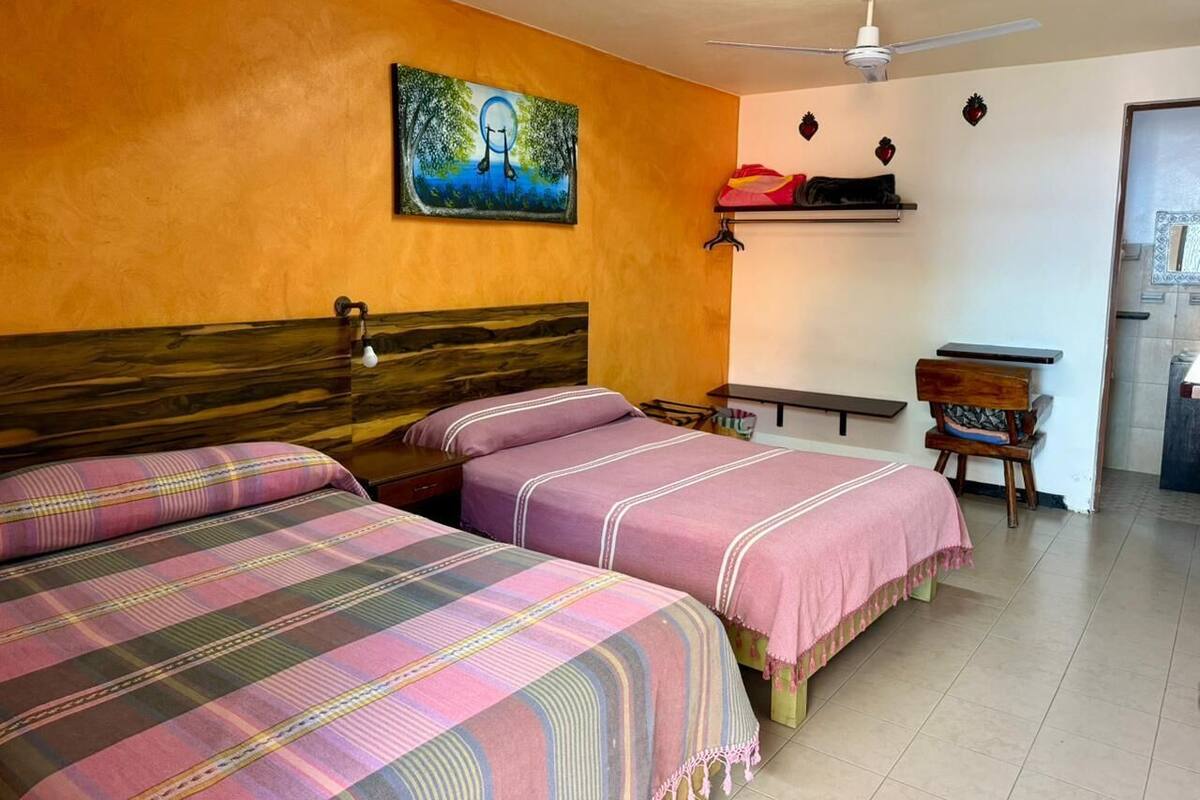 A room with two beds, a ceiling fan, a painting on the wall, and a small table with a chair.