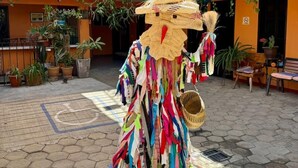 A colorful outdoor area with a person in a straw hat and a dress made of colorful ribbons. There are potted plants and a wheelchair-accessible path.