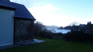 A stone building with a sloped roof, a grassy yard, and a view of a body of water and distant hills. - Shanagarry Holiday Village (Shanagarry)