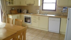 A kitchen with light-colored cabinets, a wooden dining table, chairs, a stove, a sink, and a dishwasher. - Ballylickey Bay Holiday Home (Bantry)