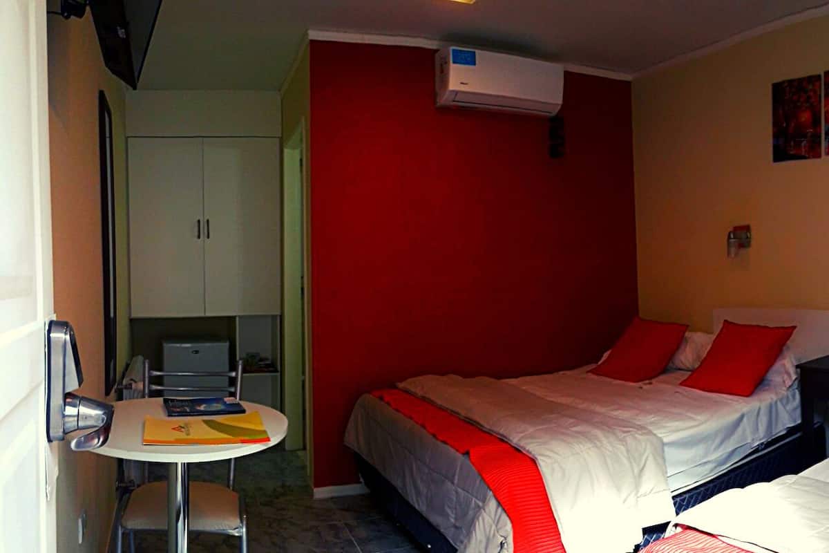 A hotel room with a bed, a desk, a chair, and a small table. There is an air conditioning unit on the wall.