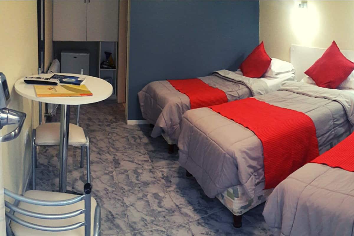 A hotel room with three beds, each with red pillows and grey bedding. There is a small round table with a chair and a few items on it. The room has a blue accent wall and a marble floor.