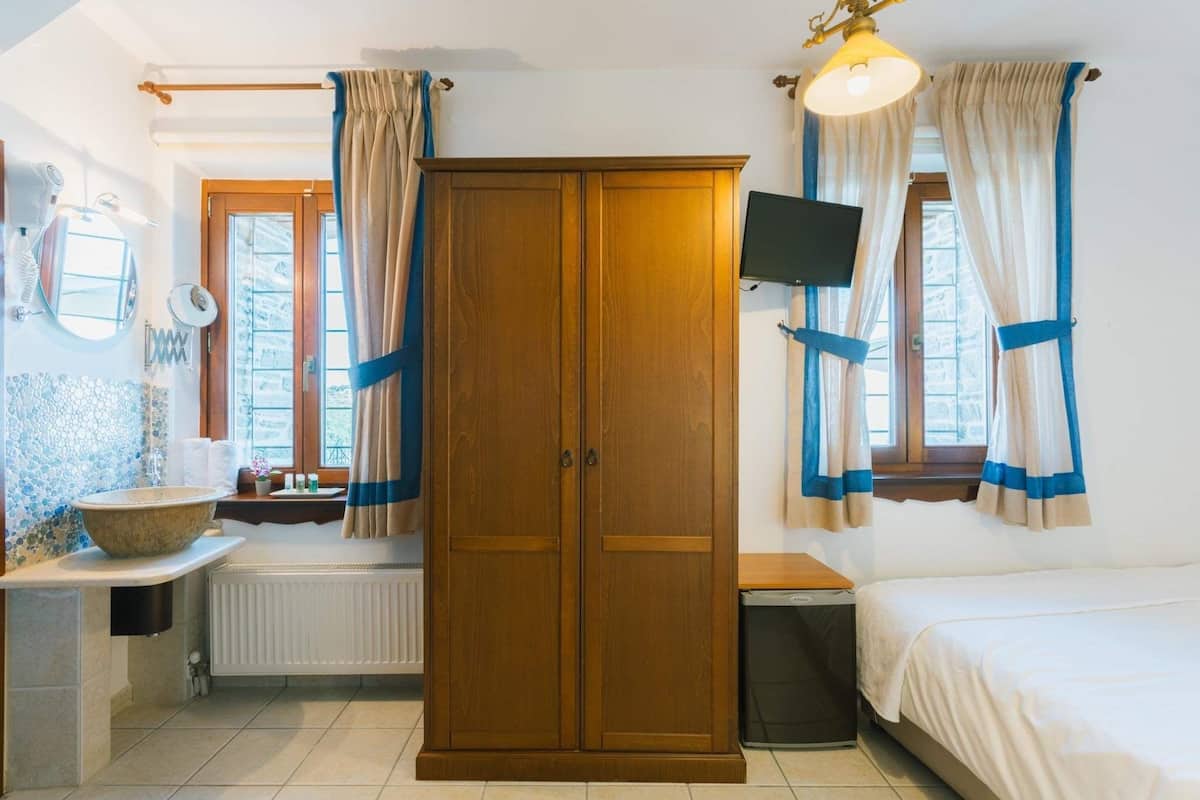 Quarto com guarda-roupa de madeira, cama, mesa de cabeceira pequena, radiador e janela com cortinas.