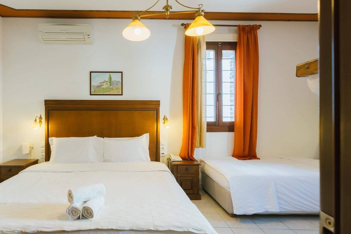 Quarto de hotel com uma cama grande, duas mesas de cabeceira, um criado-mudo, uma janela com cortinas laranja e um quadro na parede.