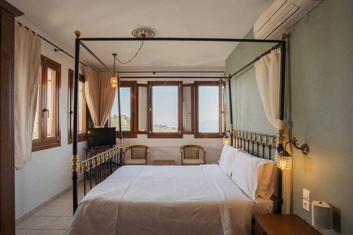 Quarto com uma cama de dossel, duas cadeiras de apoio, uma televisão e vista para o exterior.