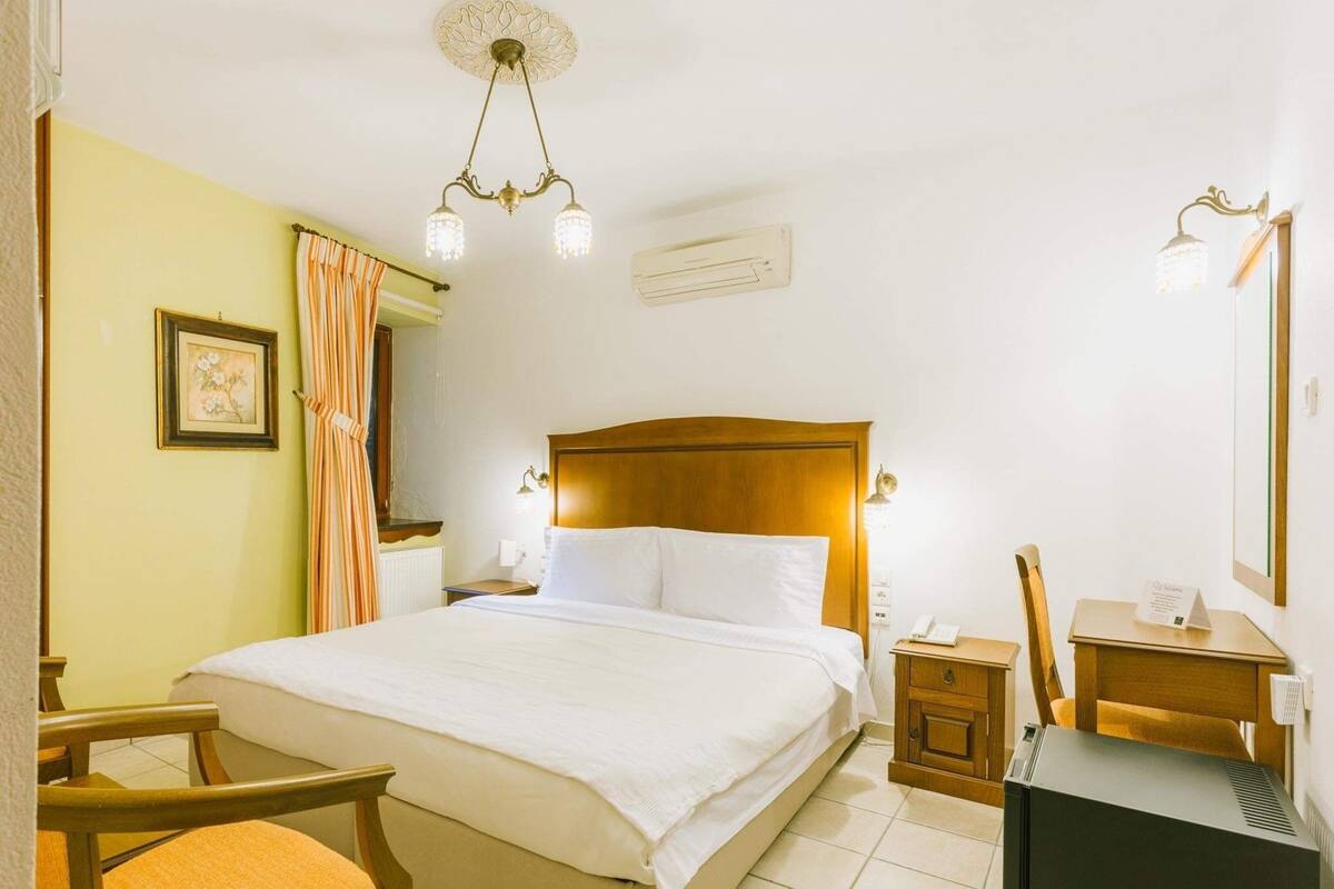 Quarto de hotel com uma cama grande, cabeceira de madeira, mesas de cabeceira, escrivaninha e uma cadeira.