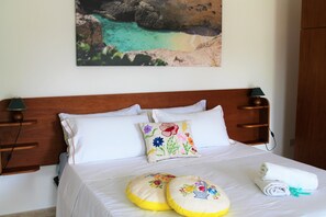 A neatly made bed with white linens, two decorative pillows, a rolled towel, and a painting of a coastal scene on the wall.