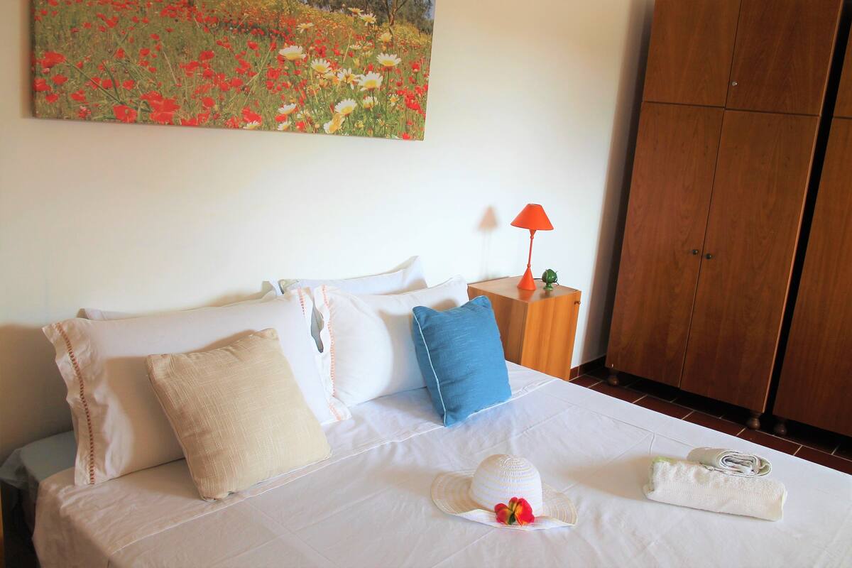 Una cama bien hecha con una colcha blanca, una almohada azul y una almohada color beige. Una lámpara roja está sobre una mesita de noche de madera junto a la cama. Un cuadro de un campo con flores rojas y amarillas cuelga en la pared sobre la cama.