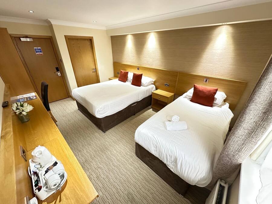 A hotel room with two beds, wooden headboards, a wooden desk with a vase of flowers, and a small table with toiletries.