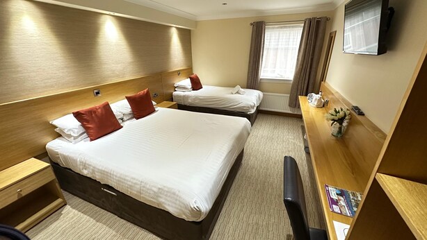 A hotel room with two beds, a desk, a chair, a TV, and a window with curtains. - Hill Park Hotel (Dunfermline)