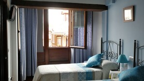 A bedroom with two beds, a window with curtains, a bedside table with a lamp, and a wall-mounted picture frame. - Pasaje San Jorge (Comillas)