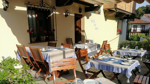 An outdoor dining area with tables set for two, wooden chairs, and a brick building in the background. - Pasaje San Jorge (Comillas)