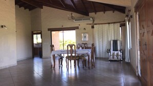 Uma ampla sala de jantar com uma mesa posta para quatro pessoas, cadeiras de madeira, um lustre e uma grande janela com cortinas.