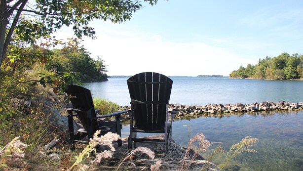 Zwei Adirondack-Stühle am Seeufer mit steiniger Küste und klarem Wasser.