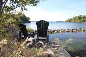 Two Adirondack chairs by a lake with a rocky shoreline and clear water.