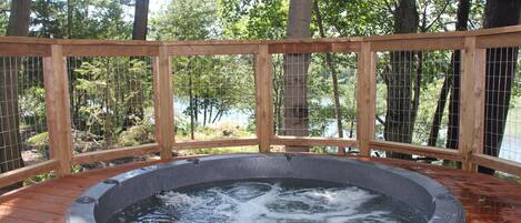 Un jacuzzi rodeado por una terraza de madera con cercado de alambre, en un área boscosa con árboles y un cuerpo de agua al fondo.