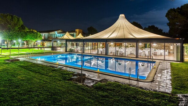 Una piscina con zona cubierta, rodeada de césped cuidado y árboles.