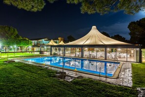 Una piscina con zona cubierta, rodeada de césped cuidado y árboles.
