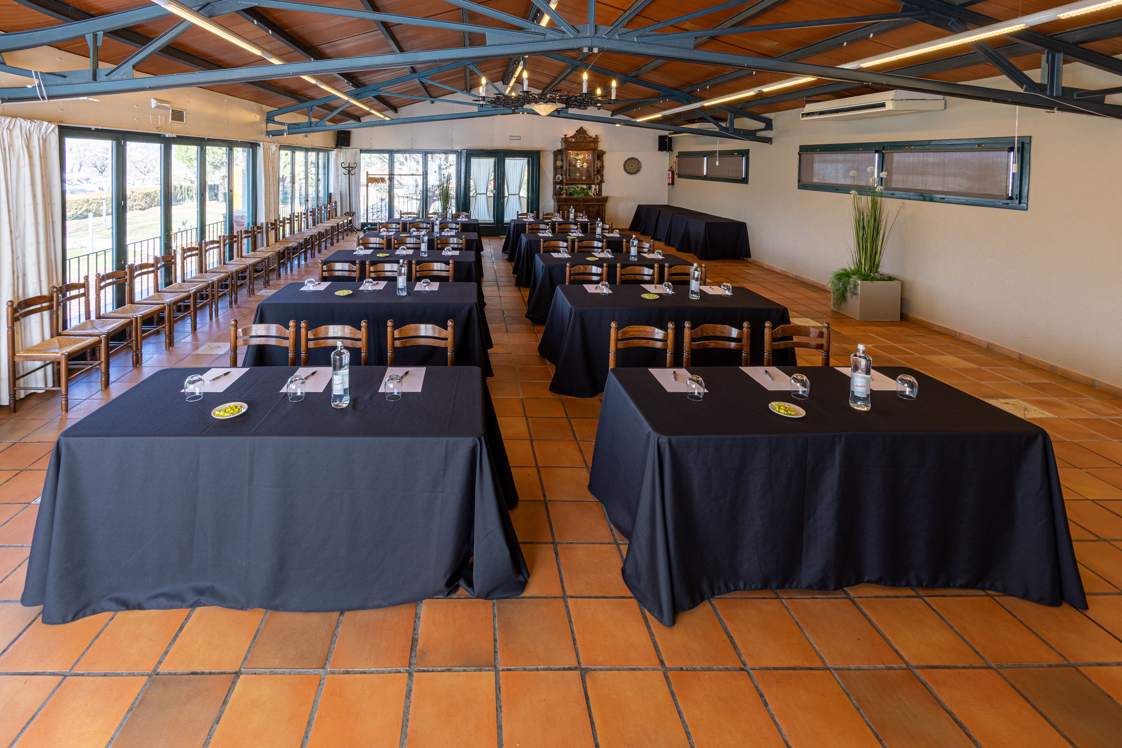 Un salón amplio con filas de mesas y sillas, ventanas grandes y un techo alto con vigas a la vista.