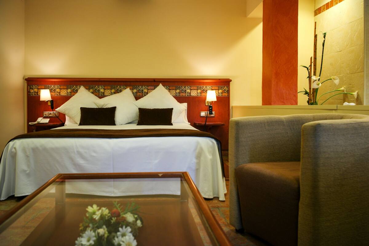 Quarto de hotel com uma cama, duas mesas de cabeceira com luminárias, uma cadeira e uma pequena mesa com um vaso de flores.