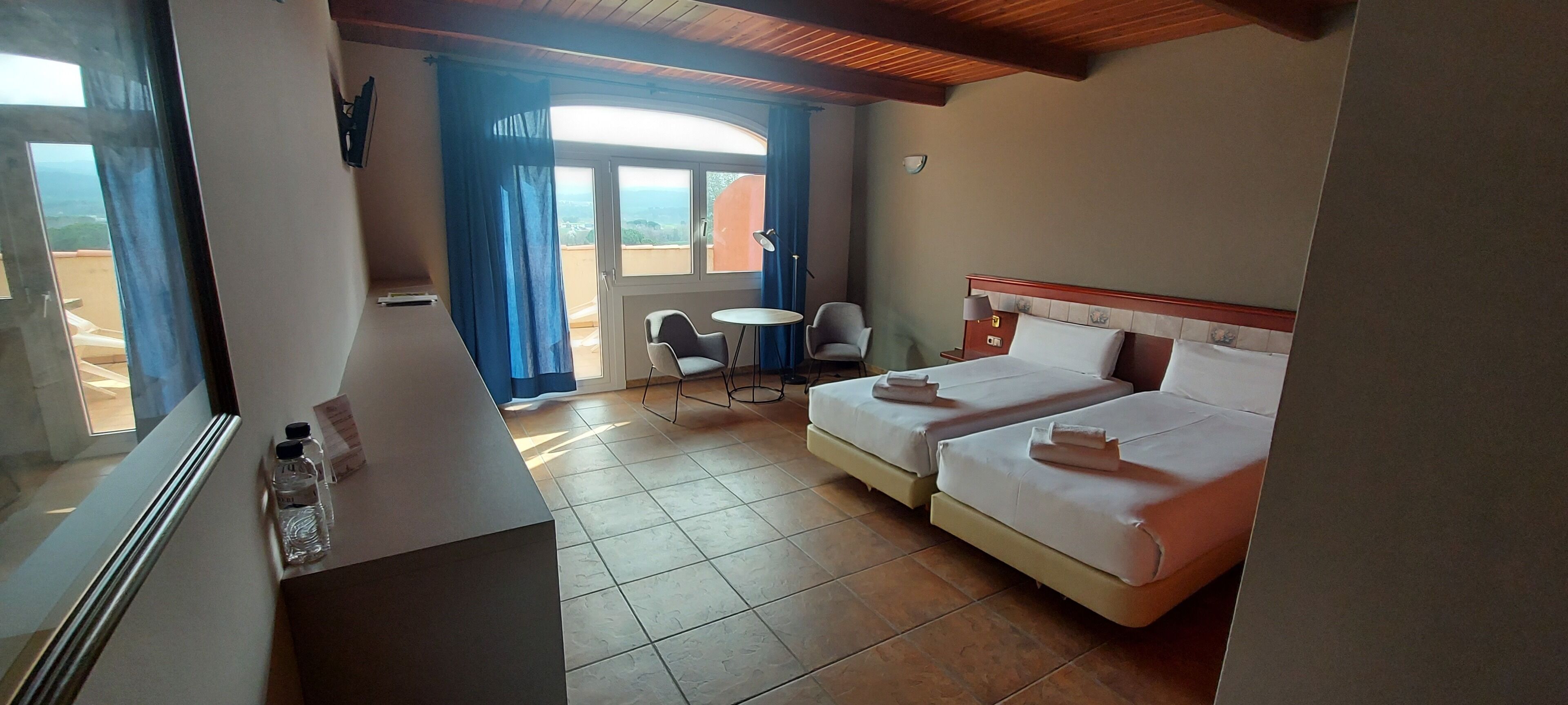 A hotel room with two beds, a small table, a chair, and a view of the outdoors.