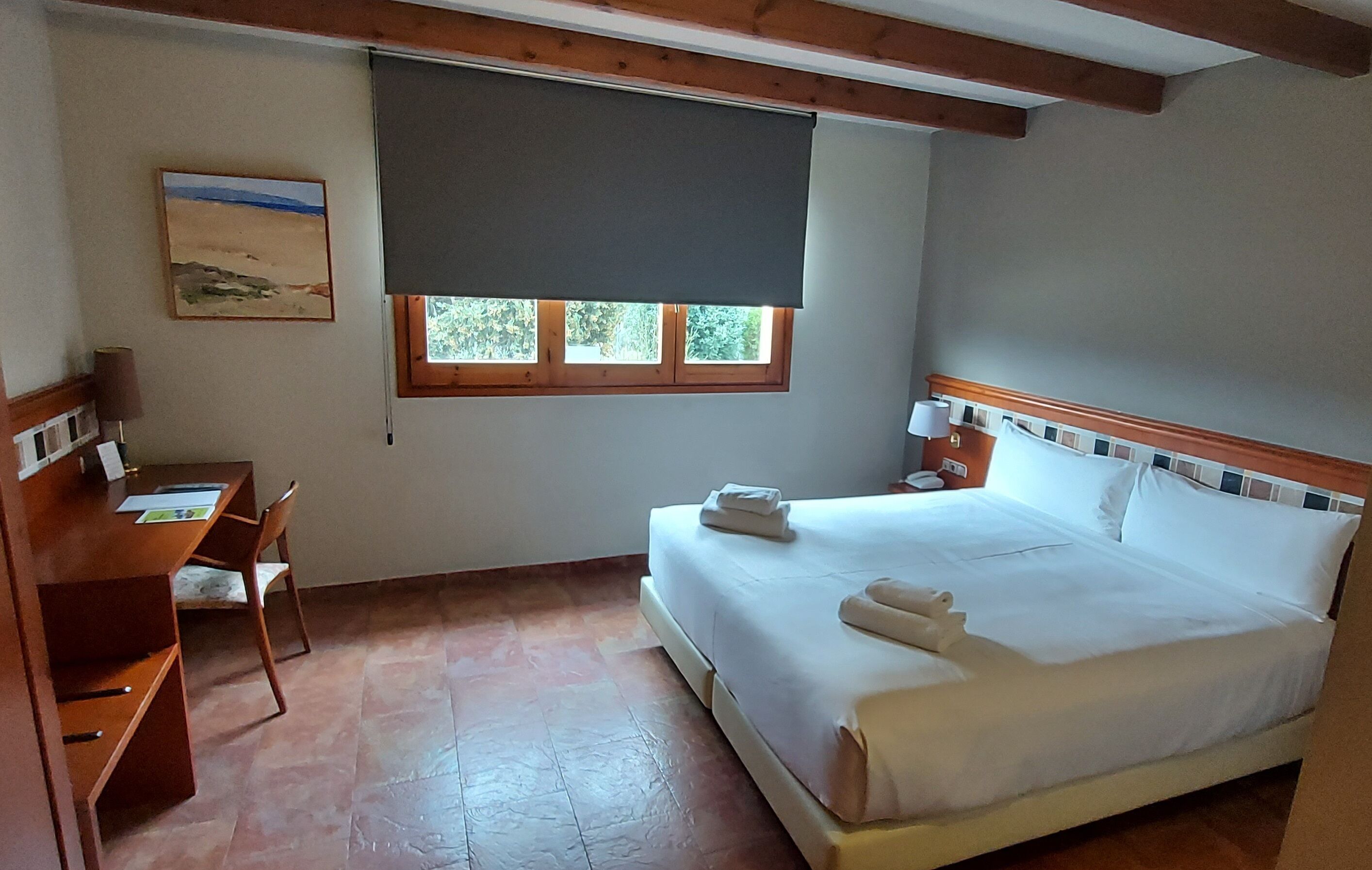 Ein Schlafzimmer mit Bett, Schreibtisch, Stuhl, Fenster mit Jalousie und einem Gemälde an der Wand.