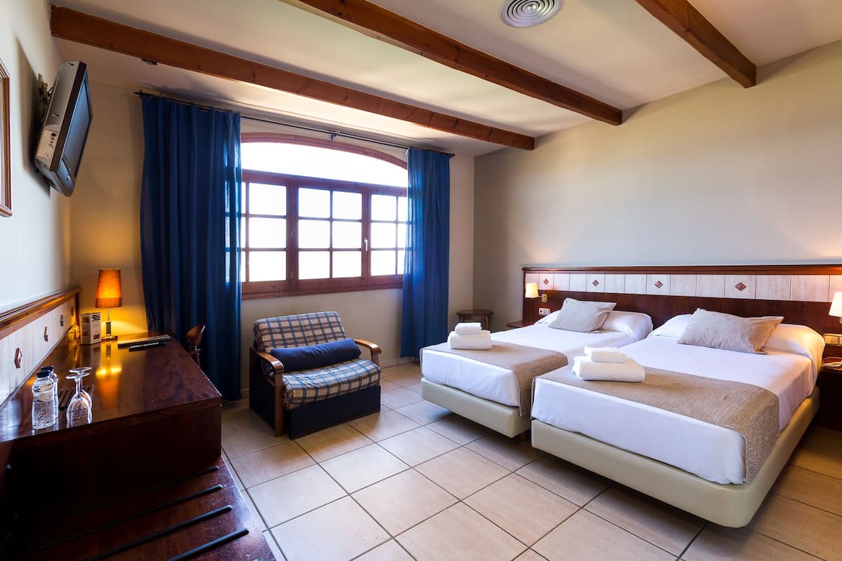 Quarto de hotel com duas camas, uma escrivaninha, uma cadeira, janela com cortinas e teto de madeira.