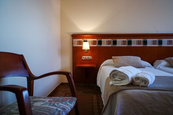 Habitación de hotel con cabecera de madera, mesita de noche con lámpara, silla con cojín floral y cama con toallas enrolladas.