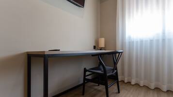 Una habitación con un escritorio de madera, una silla, un televisor montado en la pared y una ventana con cortinas traslúcidas.