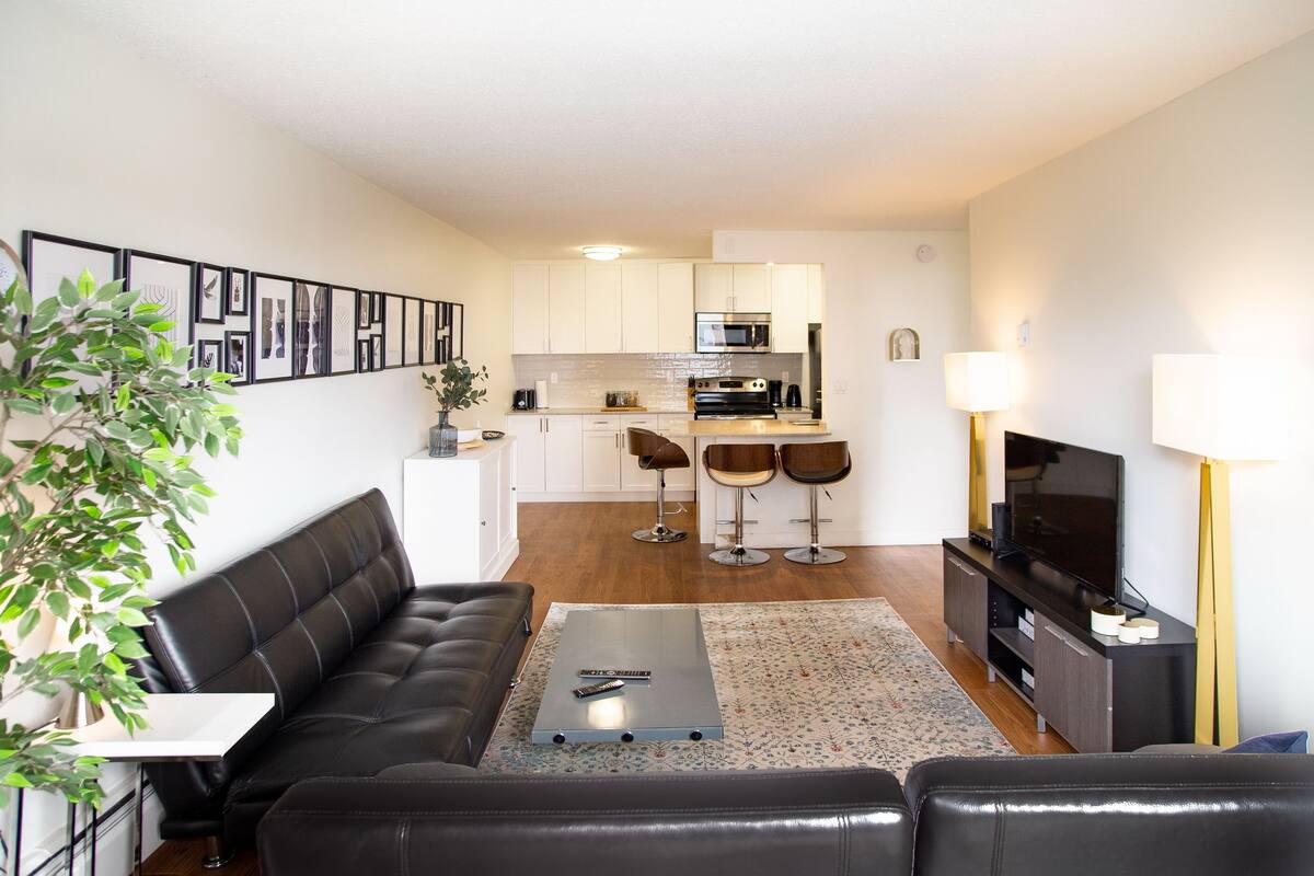 A modern living room with a black leather sofa, a coffee table, and a television. There is a kitchen in the background with white cabinets, a microwave, and a refrigerator.