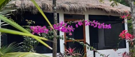 A thatched-roof structure with a white exterior, surrounded by lush greenery and pink flowers.