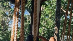 A person sitting on a swing with a white cushion, surrounded by thick ropes, in a room with a view of lush greenery. - Maya Eco Village (Akumal)