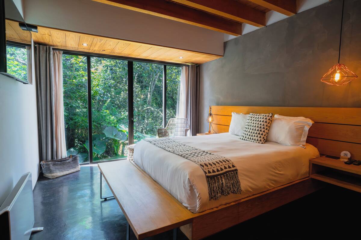 A modern bedroom with a large bed, wooden headboard, and a view of greenery through floor-to-ceiling windows.