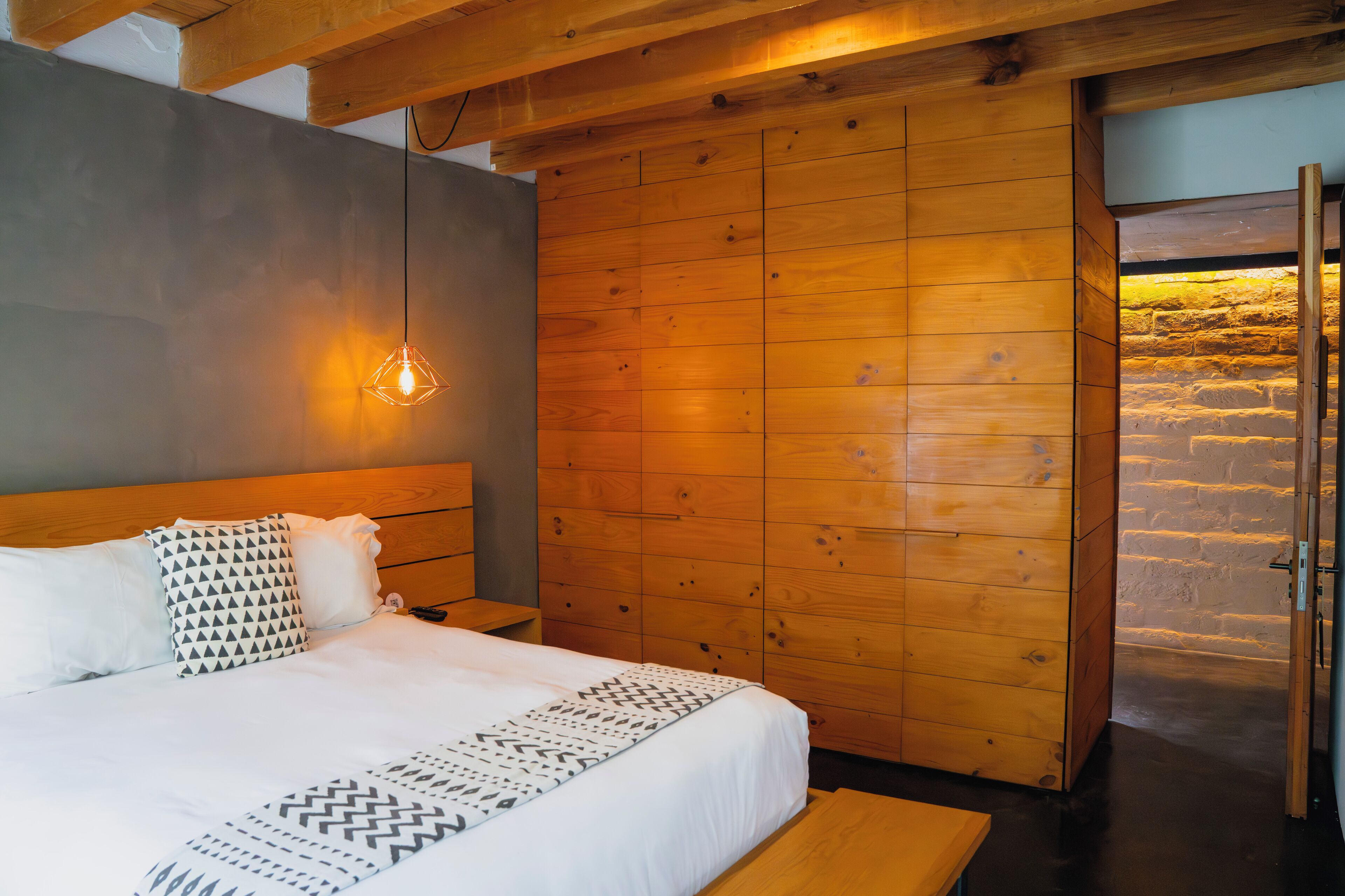 A bedroom with a wooden headboard, a bed with patterned bedding, a hanging light fixture, and a brick wall behind the bed.
