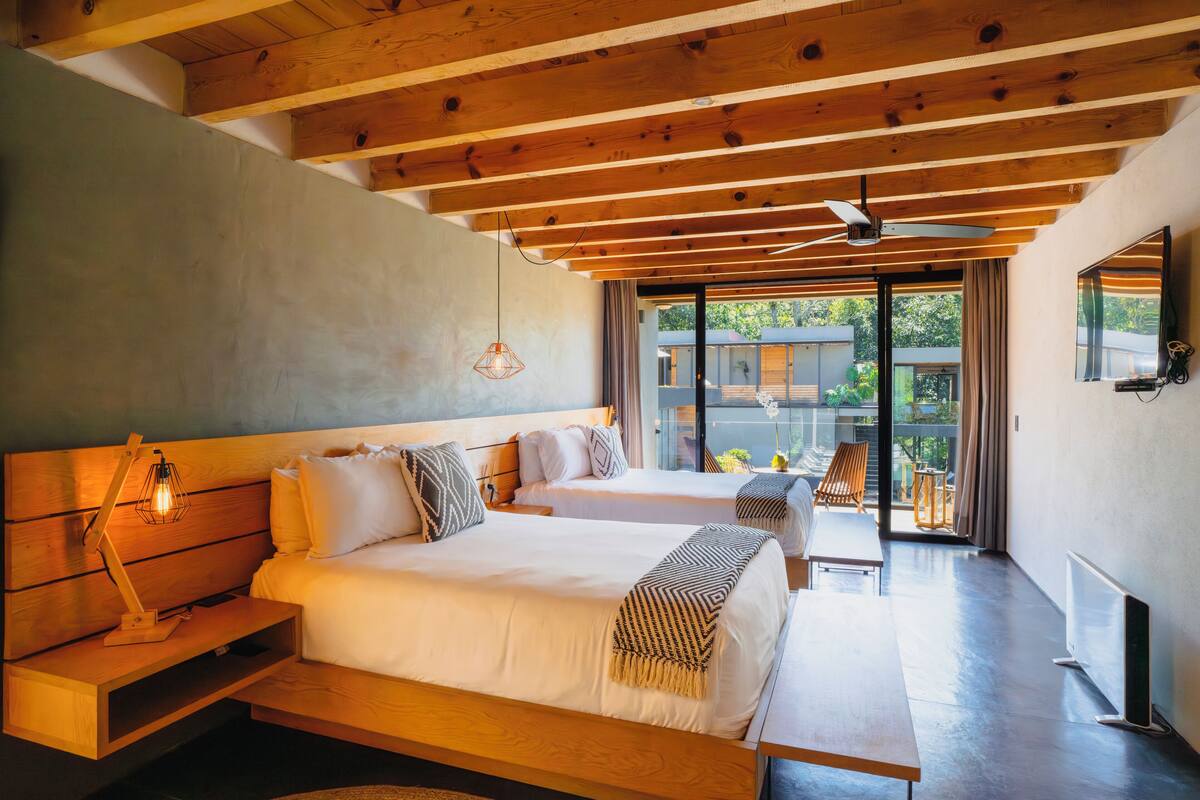 A modern bedroom with two beds, wooden beams, a ceiling fan, and a view of the outdoors.