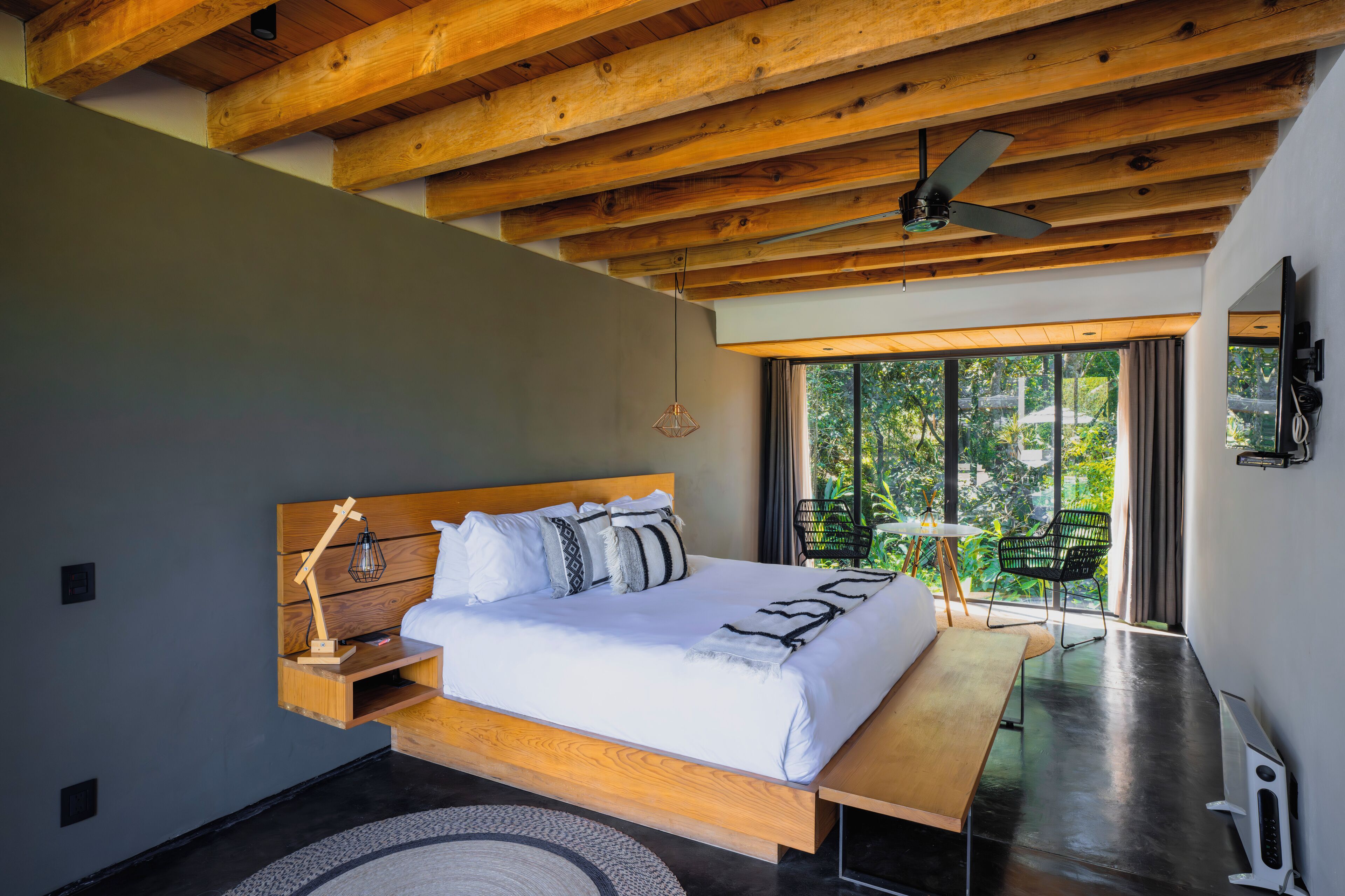 Une chambre moderne avec un plafond en bois, un grand lit doté d’une tête de lit en bois, une table de chevet avec une lampe, et une vue sur la verdure grâce à de grandes fenêtres.