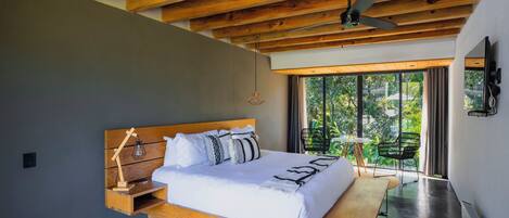 Une chambre moderne avec un plafond en bois, un grand lit doté d’une tête de lit en bois, une table de chevet avec une lampe, et une vue sur la verdure grâce à de grandes fenêtres.