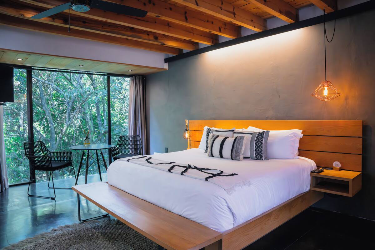 A modern bedroom with a large bed, wooden headboard, and a view of trees through large windows.