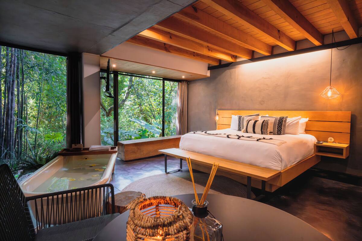 A bedroom with a large bed, a wooden headboard, a bedside table, a nightstand, a window with a view of greenery, and a bathtub.