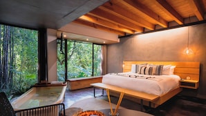 Un dormitorio con una cama grande, cabecera de madera, una mesita de noche, una ventana con vistas a la vegetación y una bañera.