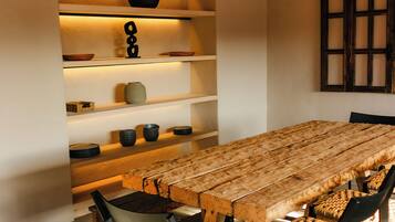 Un comedor con una mesa y sillas de madera, estantes con objetos decorativos y una ventana con diseño de cuadrícula.