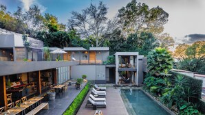 Un espacio moderno para comer al aire libre con mesas y bancos de madera, una piscina y abundante vegetación.