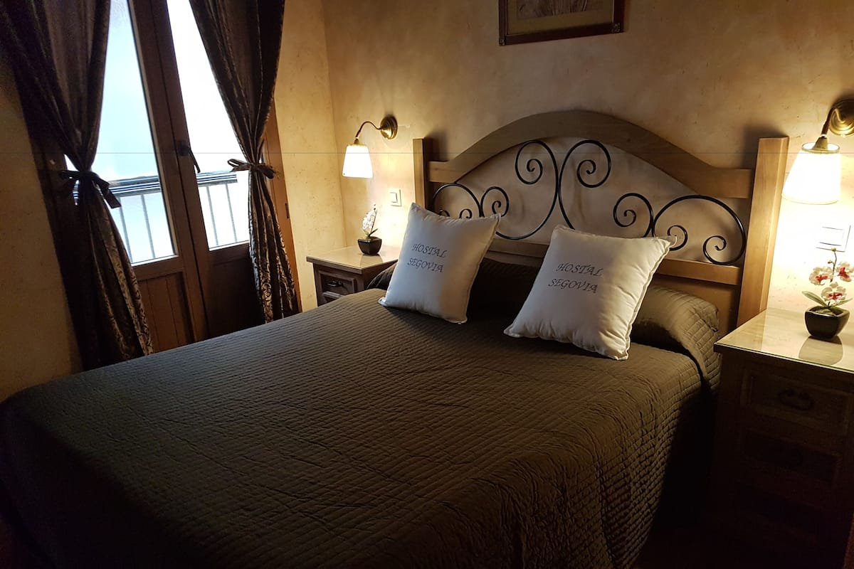 A neatly made bed with two pillows, a headboard with a decorative design, bedside lamps, and a small table with a plant.