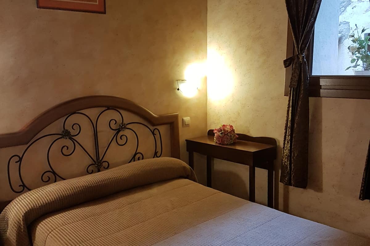 A bedroom with a bed, a nightstand, a wall-mounted light, a framed picture, and a window with curtains.