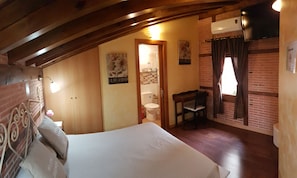 A bedroom with a wooden ceiling, a bed, a small table, a chair, a window with curtains, and a bathroom visible through an open door. - Hostal Segovia (Segovia)