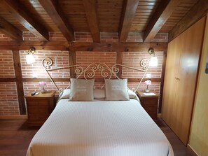 A bedroom with a wooden ceiling, brick walls, a bed with white bedding, two bedside tables with lamps, and a wooden wardrobe. - Hostal Segovia (Segovia)