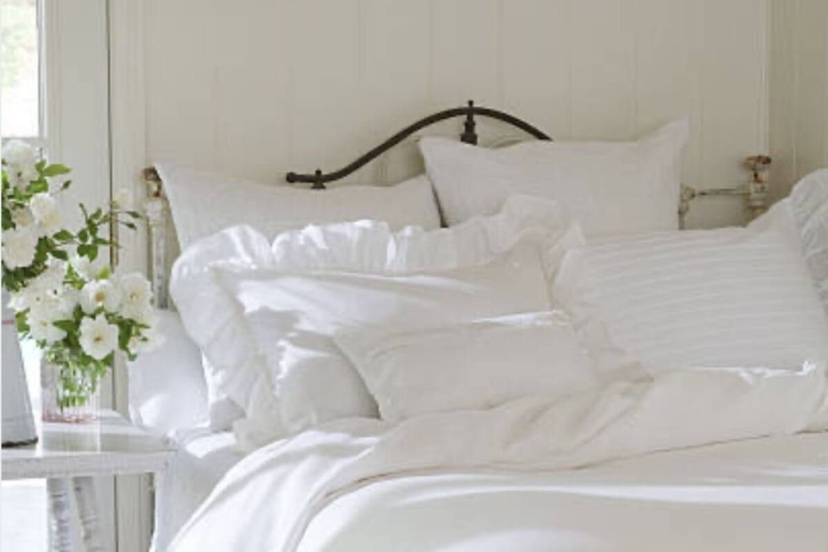 A neatly made bed with white bedding and a ruffled skirt, a bedside table with a vase of flowers, and a window with blinds.