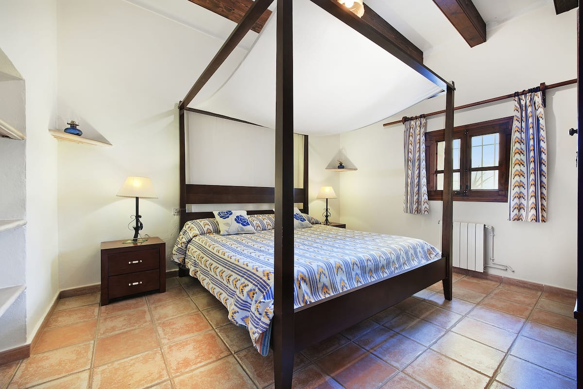 A four-poster bed with a patterned bedspread, a nightstand with a lamp, and a window with curtains.