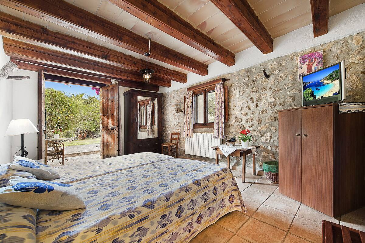 A bedroom with a bed, wooden beams, a stone wall, a television, and a view of the outdoors.